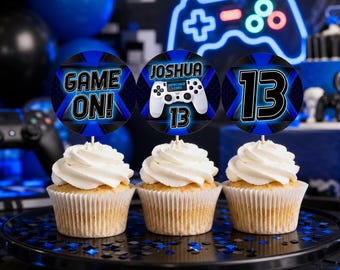 May include: Three cupcakes with white frosting, each topped with a circular blue and black gaming-themed topper. The toppers feature the text "GAME ON!", the name "JOSHUA" with a game controller graphic and the number "13", and the number "13".