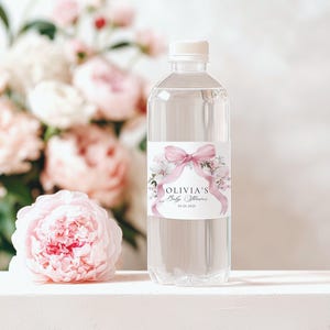 May include: A clear plastic water bottle with a white cap and a label that reads "Olivia's Baby Shower" with a pink bow and floral design. A pink peony and a bouquet of pink and white flowers are in the background.
