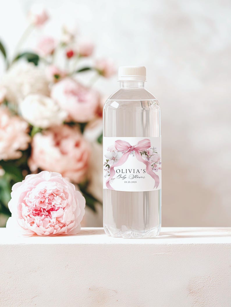 May include: A clear plastic water bottle with a white cap and a label that reads "Olivia's Baby Shower" with a pink bow and floral design. A pink peony and a bouquet of pink and white flowers are in the background.