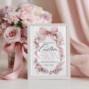 May include: A framed sign featuring a floral wreath and a pink bow. The sign says "Custom Sign" and "Add Your Own Text" with an 8x10 inch size. A bouquet of pink and purple roses is in the background.