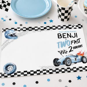Può includere: Allestimento tavola per una festa di compleanno a tema corse. Include una tovaglietta con un'illustrazione di auto da corsa e il testo "Benji is Two Fast 2 Curious". Il tavolo ha anche un piatto blu, una forchetta e un cupcake.
