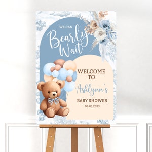 May include: A blue and beige baby shower welcome sign with the text "We can Bearly Wait" and "Welcome to Ashlynn's Baby Shower 06.03.2023". The sign features a teddy bear holding blue and brown balloons.