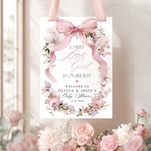 May include: A baby shower sign with a pink ribbon and floral design. The sign reads "A Sweet Little Girl Is On Her Way!" with the names Olivia & Jhon and the date 10.23.2025. The sign is surrounded by pink and white flowers.