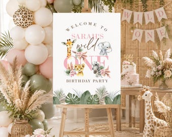 May include: A white rectangular birthday party welcome sign on a wooden easel featuring the text 'WELCOME TO SARAH'S wild ONE BIRTHDAY PARTY' with illustrations of a giraffe, elephant, lion, and zebra surrounded by pink flowers and green palm leaves.