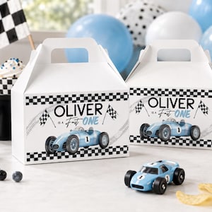 May include: White gable boxes with black and white checkered borders and racing car graphics. The boxes read "Oliver is a Fast One." A blue toy race car sits in front of the boxes, with cupcakes and balloons in the background.