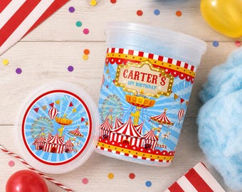 Pode incluir: Um recipiente e tampa de plástico transparente com um design temático de circo. O recipiente apresenta um fundo azul com uma roda gigante, tendas e o texto "CARTER'S 1ST BIRTHDAY". Detalhes em vermelho, branco e azul são visíveis.