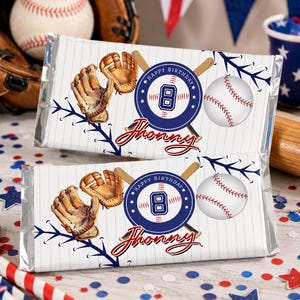 May include: Two baseball-themed candy bars with white and blue pinstripes. Each wrapper features a baseball, glove, and the text "Happy Birthday" with the name "Jhonny". A baseball glove and ball are in the background.