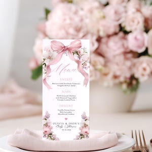 May include: A baby shower menu card featuring a pink bow and floral decorations. The menu lists entree, main, and dessert choices. The card rests on a white plate with a pink napkin, with a bouquet of pink flowers in the background.