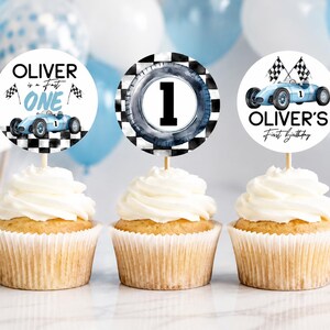 May include: Three cupcakes with white frosting and decorative toppers. The toppers feature racing car designs, checkered flags, and the text "Oliver is a Fast One" and "Oliver's First Birthday". The cupcakes are set against a backdrop of balloons and checkered flags.