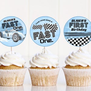 Può includere: Tre cupcake con glassa bianca, ciascuno sormontato da una decorazione circolare azzurra. Le decorazioni presentano temi di corse, tra cui un'auto da corsa d'epoca, bandiere a scacchi e il testo "Oliver's Fast One" e "Oliver's First Birthday".