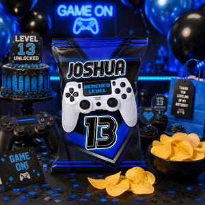 Op de afbeelding: Een gepersonaliseerde zak chips met een blauw en zwart geometrisch ontwerp, de naam JOSHUA, een witte gamecontroller-afbeelding met de tekst REACHED LEVEL en het getal 13. De zak is omringd door feestdecoraties zoals een taart en snacks.