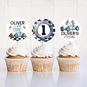 Può includere: Tre cupcake con glassa bianca sormontati da decorazioni rotonde di carta per cupcake. Le decorazioni raffigurano un'auto da corsa blu con bandiere a scacchi, un cerchio a scacchi bianco e nero con il numero uno e un cerchio bianco con un'auto da corsa blu e il testo "Oliver's First Birthday".
