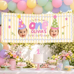 Op de afbeelding: Een rechthoekige verjaardagsbanner met gele en witte verticale strepen, voorzien van de tekst "it's fun to be one OLIVIA'S first birthday" in kleurrijke letters, twee feesthoedjes en twee ronde foto's van een babygezicht.