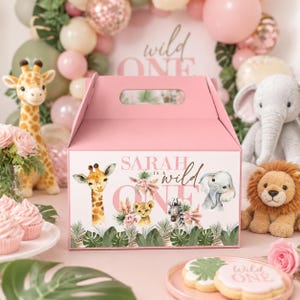 Op de afbeelding: Een roze kartonnen doos met handvat, bedrukt met een giraffe, leeuwenwelp, zebra en olifant tussen tropische bladeren. De tekst Sarah is a wild ONE staat op het voorpaneel in een combinatie van schreefletters en een handschriftlettertype.