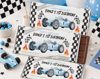 May include: Three chocolate bars with racing car designs, checkered flag accents, and the text "HAPPY BIRTHDAY BENJI'S 1ST BIRTHDAY". A toy race car and checkered flags are also present. Blue and white balloons and candies complete the birthday theme.