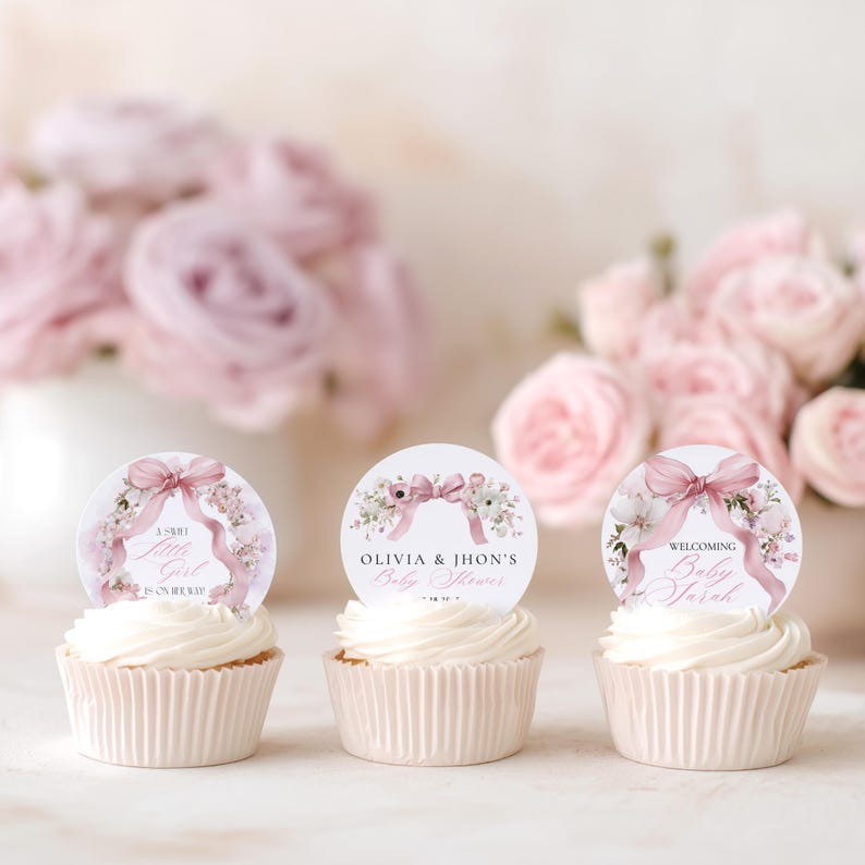 May include: Three cupcakes with white icing and decorative toppers. The toppers feature floral designs, pink bows, and text that reads "A Sweet Little Girl is on her way!", "Olivia & Jhon's Baby Shower", and "Welcoming Baby Sarah".