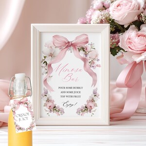 May include: A white framed sign reading "Mimosa Bar" with a pink bow and floral decorations. A bottle of orange juice with a floral tag is in front. The sign says: "Pour some bubbly, add some juice, top with fruit, enjoy!"