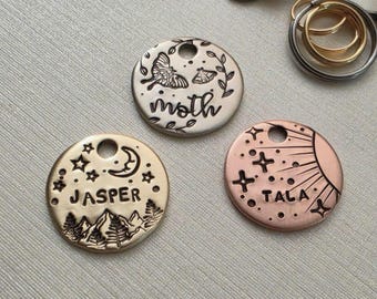 May include: Three circular metal pet ID tags with engraved designs. One silver tag features a moth and the word moth. One gold tag shows mountains, stars, and the name Jasper. One copper tag displays a sun, stars, and the name Tala.