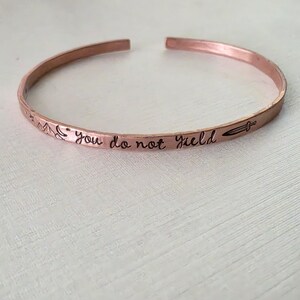 Könnte beinhalten: Ein Kupfer-Armreif mit gehämmertem Finish und offenem Design. Die Außenseite ist mit dem Text "you do not yield" in Schreibschrift graviert, ergänzt durch kleine eingravierte Symbole eines Gebirges, eines Sterns und eines Schwertes.