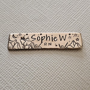 May include: A rectangular metal name tag with the name "Sophie W" and "RN" engraved. The tag features a mountain and deer design, along with stars and a crescent moon. The tag is silver in color.