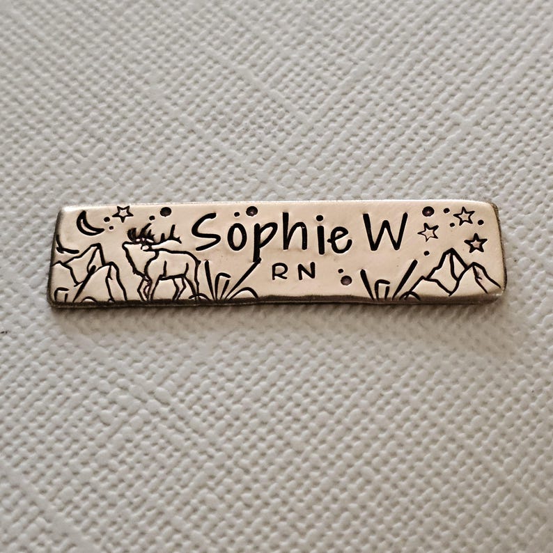 May include: A rectangular metal name tag with the name "Sophie W" and "RN" engraved. The tag features a mountain and deer design, along with stars and a crescent moon. The tag is silver in color.