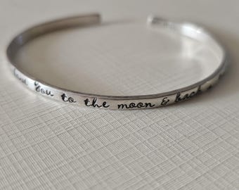 May include: A silver cuff bracelet with the inscription "I love you to the moon & back" in a handwritten style font. The bracelet is open and has a smooth, polished finish. The bracelet is resting on a textured white surface.