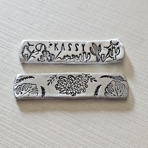 May include: Two silver metal bars with etched designs. The top bar features the name "KASSY" and desert-themed illustrations. The bottom bar showcases a floral design with leaves. Both bars have a rectangular shape with rounded edges.