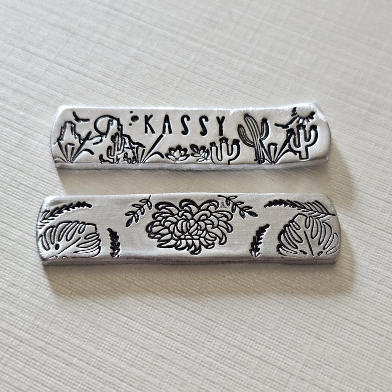 May include: Two silver metal bars with etched designs. The top bar features the name "KASSY" and desert-themed illustrations. The bottom bar showcases a floral design with leaves. Both bars have a rectangular shape with rounded edges.