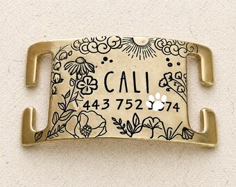 May include: A gold-colored pet tag with the name "CALI" and a phone number. The tag is decorated with black floral and cloud designs, a sun, and a paw print.
