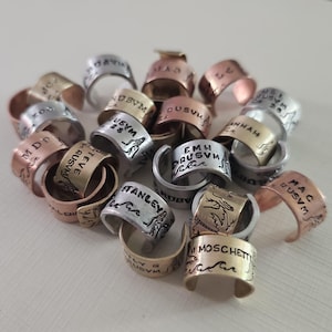 May include: A collection of handcrafted metal rings in various colors, including silver, gold, and copper. Each ring features engraved text and decorative designs. The rings are stacked together, showcasing their unique details.