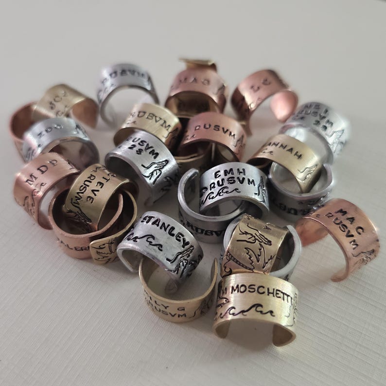 May include: A collection of handcrafted metal rings in various colors, including silver, gold, and copper. Each ring features engraved text and decorative designs. The rings are stacked together, showcasing their unique details.