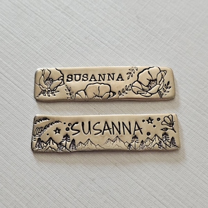 May include: Two rectangular metal name tags with the name "SUSANNA" engraved in the center. One tag features floral and bear designs, while the other showcases a mountain and forest scene. The tags are silver in color.