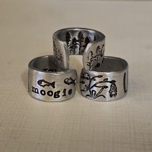May include: Three silver-colored, adjustable rings. One ring features the word "moogie" with fish illustrations. Another ring has a forest scene, and the third ring has a botanical design with a bat. The rings are stacked.