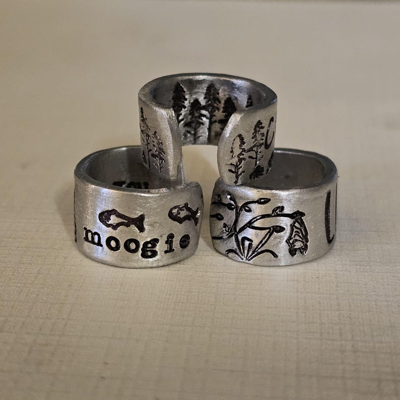 May include: Three silver-colored, adjustable rings. One ring features the word "moogie" with fish illustrations. Another ring has a forest scene, and the third ring has a botanical design with a bat. The rings are stacked.