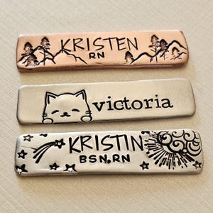 May include: Three rectangular metal name tags. The top tag is copper-colored with the name "KRISTEN RN" and a mountain and tree design. The middle tag is silver with the name "victoria" and a cat face. The bottom tag is silver with the name "KRISTIN BSN, RN" and a sun and star design.