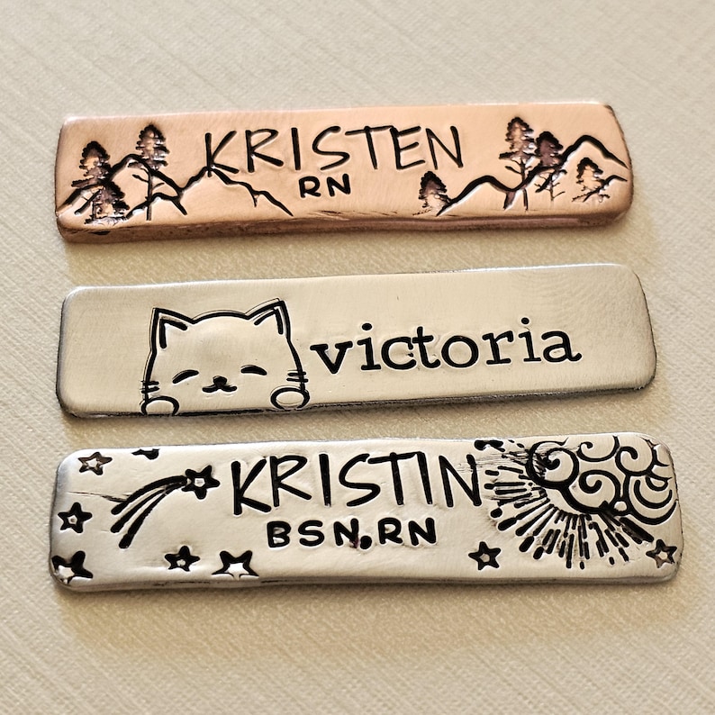 May include: Three rectangular metal name tags. The top tag is copper-colored with the name "KRISTEN RN" and a mountain and tree design. The middle tag is silver with the name "victoria" and a cat face. The bottom tag is silver with the name "KRISTIN BSN, RN" and a sun and star design.