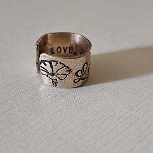 May include: A bronze-colored ring with the word "LOVE" engraved on the top. The ring features a floral design and the word "Love" in script. The ring is a unique piece of jewelry.