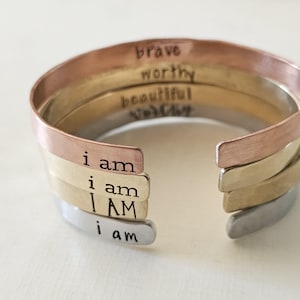Op de afbeelding: Een set van vier gestapelde armbanden van koper, messing en zilver met de woorden "brave", "waardig", "mooi" en "ik ben" erop gegraveerd.