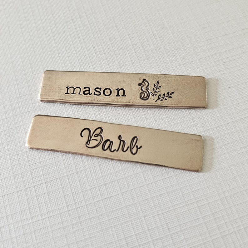 May include: Two rectangular metal name tags. One tag reads "mason" with a seahorse and leaf design. The other tag reads "Barb" in cursive. Both tags have a brushed metal finish and are set against a textured white background.