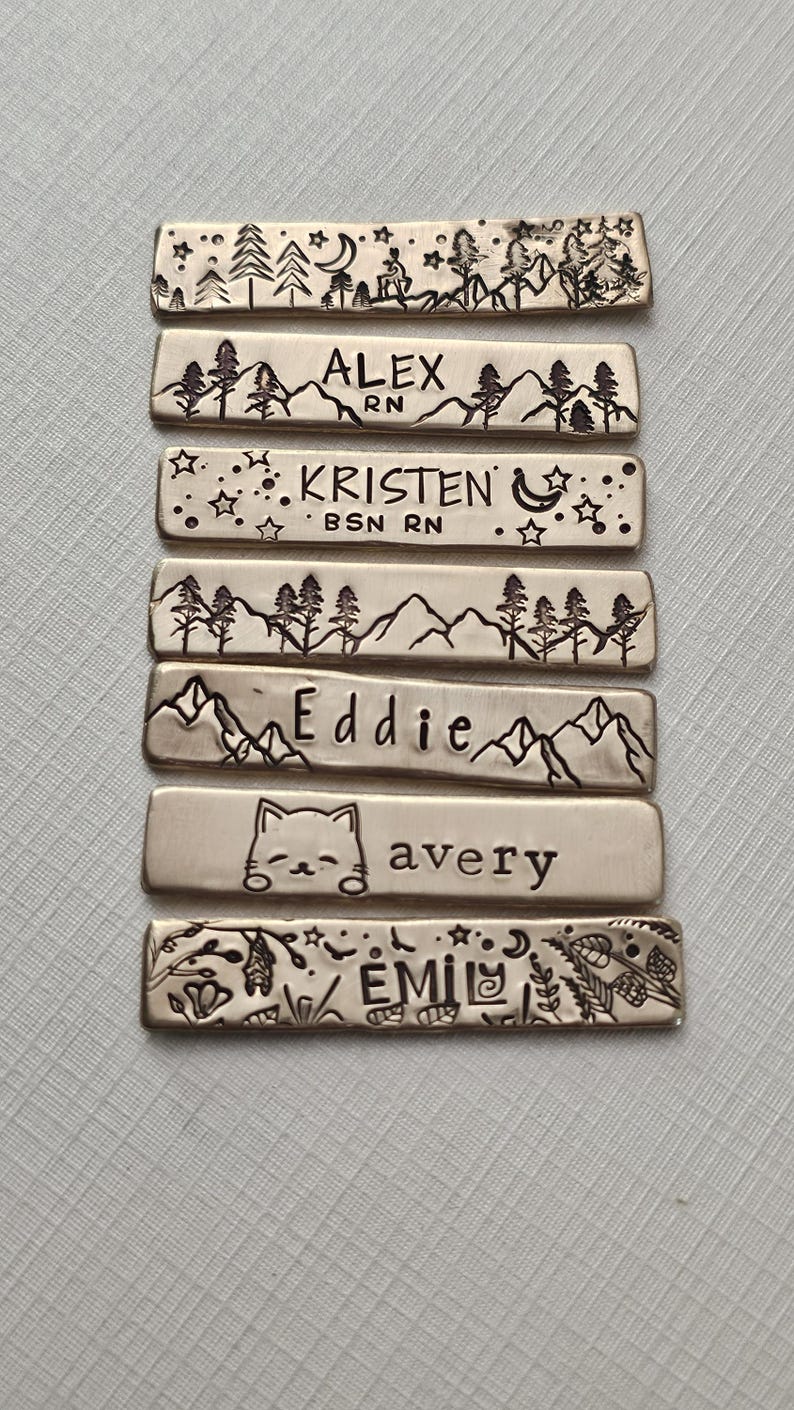 May include: A collection of rectangular metal tags with engraved designs and names. Each tag features a unique landscape or animal illustration, along with a name. Some tags include the text "RN" and "BSN RN".