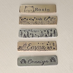 May include: A collection of rectangular metal pet tags with stamped designs and names. The tags feature a cat, fish, a forest scene with a deer, a sunburst, and paw prints. Names include Rosie, Riley, Emma, and Conniya.