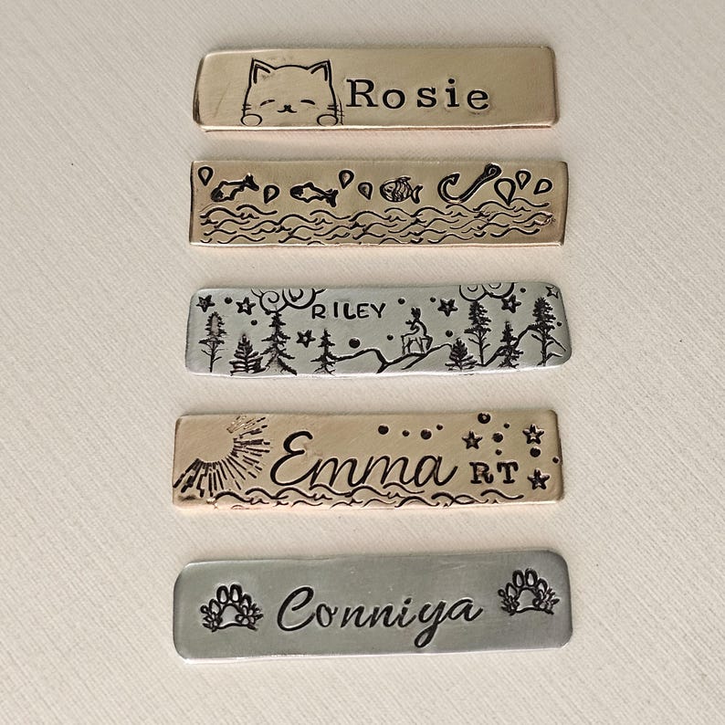 May include: A collection of rectangular metal pet tags with stamped designs and names. The tags feature a cat, fish, a forest scene with a deer, a sunburst, and paw prints. Names include Rosie, Riley, Emma, and Conniya.