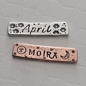 May include: Two metal pet tags. The top tag is silver with the name "April" and paw prints. The bottom tag is copper-colored with the name "Moira", stars, and a crescent moon.