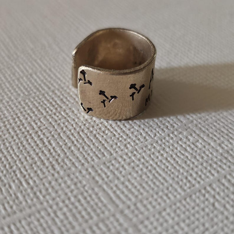 May include: A gold-colored ring with a wide band and an open design. The ring features a series of small, dark-colored, stylized symbols etched around the band. The ring is set against a textured, off-white surface.