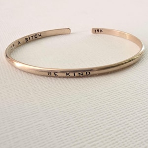May include: A thin, gold-toned metal cuff bracelet with an open design. The exterior is stamped with the text BE KIND, while the interior features the stamped text OF A BITCH and the marking 14K. The bracelet has a smooth, polished finish.