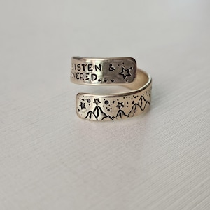 Può includere: Anello avvolgente color argento con dettagli incisi. L'anello presenta le parole "LISTEN & ANSWERED" e disegni di stelle e montagne. L'anello è progettato per avvolgersi attorno al dito.
