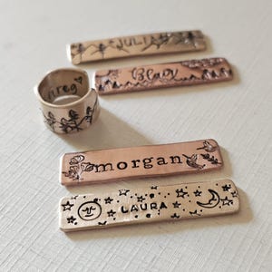 May include: A collection of personalized metal jewelry pieces. Includes a silver ring with the name "Greg" and a landscape design, and four rectangular metal tags with names and decorative elements like stars, flowers, and birds. The tags are in copper and bronze tones.