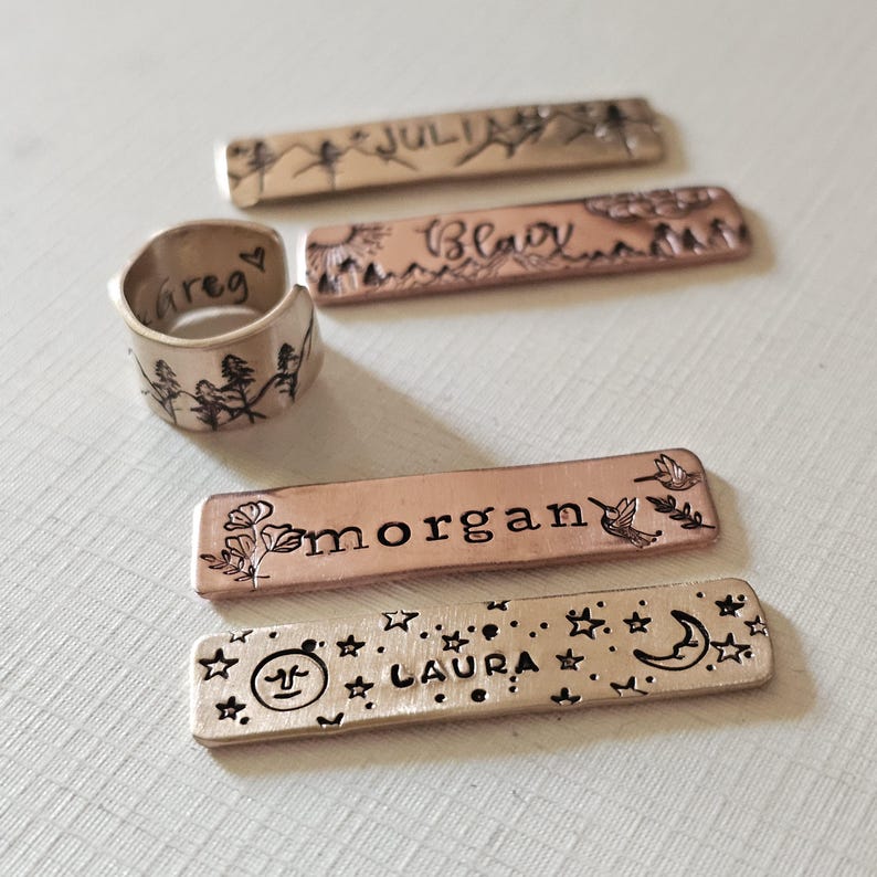May include: A collection of personalized metal jewelry pieces. Includes a silver ring with the name "Greg" and a landscape design, and four rectangular metal tags with names and decorative elements like stars, flowers, and birds. The tags are in copper and bronze tones.