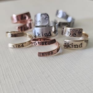 Puede incluir: Una colección de anillos de plata y cobre con las palabras "be kind" grabadas. Los anillos están apilados y dispuestos sobre una superficie blanca.