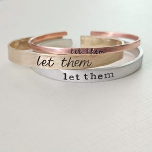 Könnte beinhalten: Drei Metallarmbänder übereinander gestapelt. Das obere Armband ist kupferfarben, das mittlere goldfarben und das untere silberfarben. Auf jedem Armband steht "let them" eingraviert.